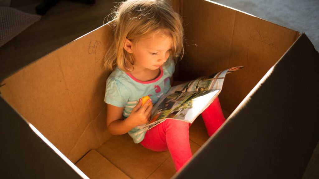Criança lendo dentro de caixa de papelão.